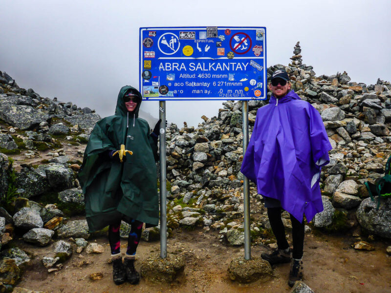 A woman and man wearing ponchos posing in the rain next to a sign for Salkantay Pass, the highest point on the Salkantay Trail. Piled rocks and clouds behind them.