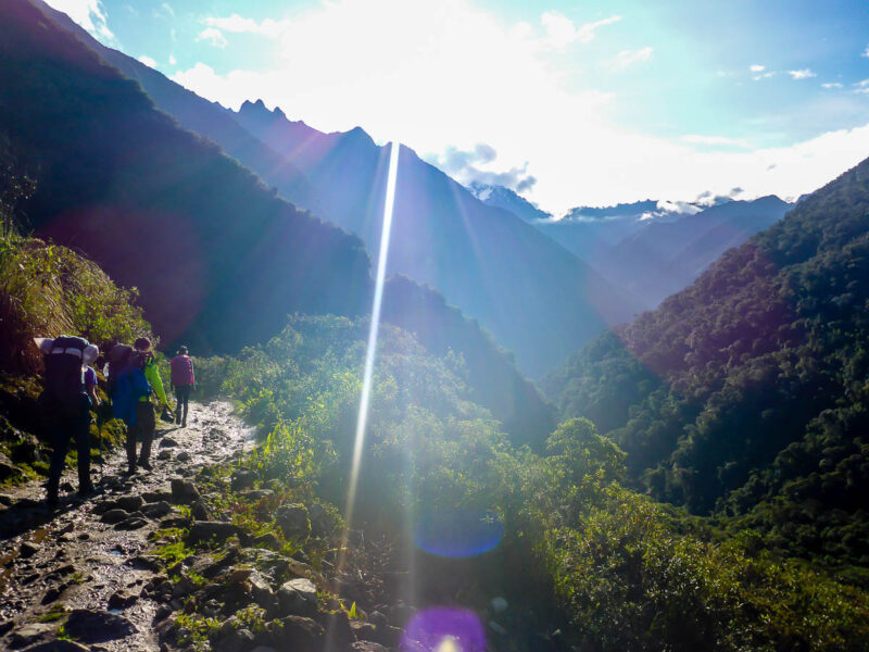 A few people hiking along a muddy, rocky trail in the Peruvian Andes with lush green plants, trees, and greenery covered mountains. The sun is bright and shining with blue skies and a few clouds.