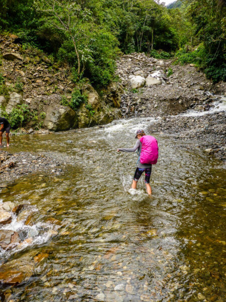 A woman wading across a river completely covering a road. Flowing river and rocks with rocky mountain in the background.