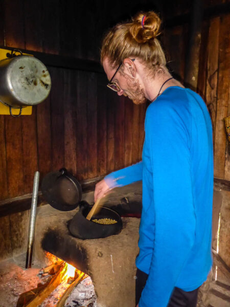 A man stirring a large pot, roasting coffee beans over an open-flame in a small, dark wooden hut.