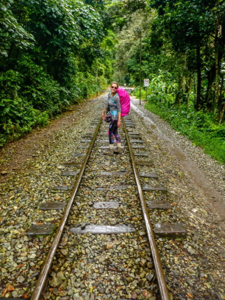 A woman with a large pink backpack, smiling at the camera as she walks along train tracks overtop a rocky trail with green trees on either side.