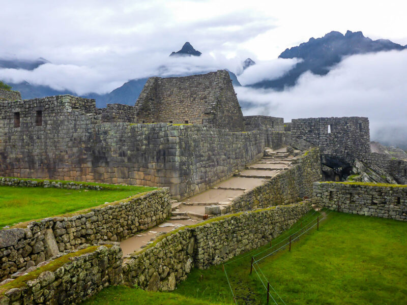 Machu Picchu grounds with Inca-built roads, walls, homes and other structures. A view of Machu Picchu mountain in the background and low-handing clouds.