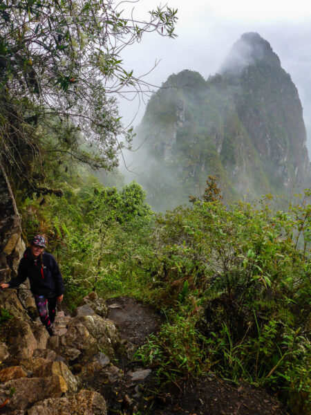 A woman posing on a steep trek on a narrow part of the trail up a mountain high up in the Andean mountains in Peru. The weather is cloudy and rainy and the jungle is lush and green.