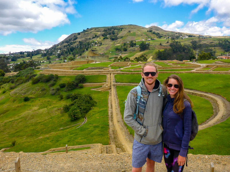 A man and woman posing in front of Ingapirca Ruins in Ecuador. There is lush bright green grass throughout the ruins and on the mountain in the background. The ruins are light beige colored stones piled to form structures.