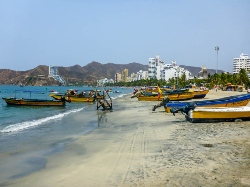 White sand beach with small colorful fishing boats in the sand and the water. White cityscape in the background with tall buildings and mountains behind it. 