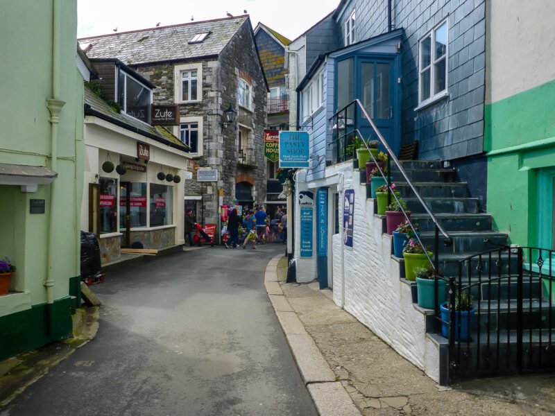 Southwest English street with light blue colored pasty shop on white side and steps on the side of it with flowers on each step. Shops line both sides of the streets in muted pastel colors, and old stones.