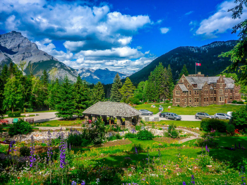 Two brick buildings set amidst green forest, purple and red flowers, gardens, forests, mountains and snow capped hills.