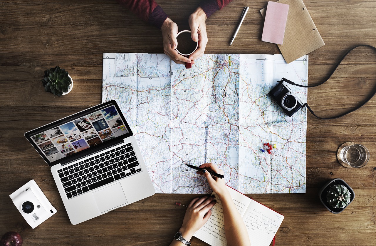 Aerial view of two people planning a trip with a map, laptop, notebook, and camera.