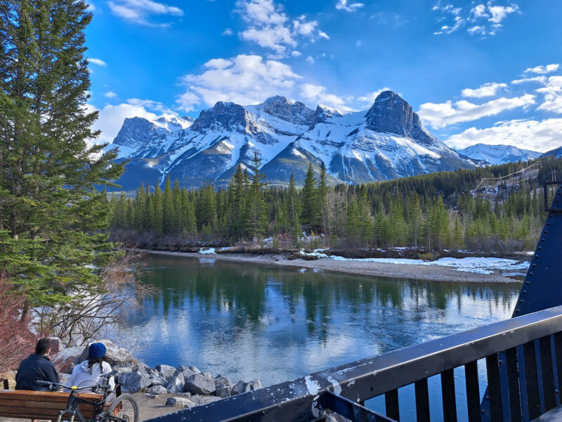 A view of snow capped Rocky Mountains with green forests, glacier water, and two cyclists sitting on a bench