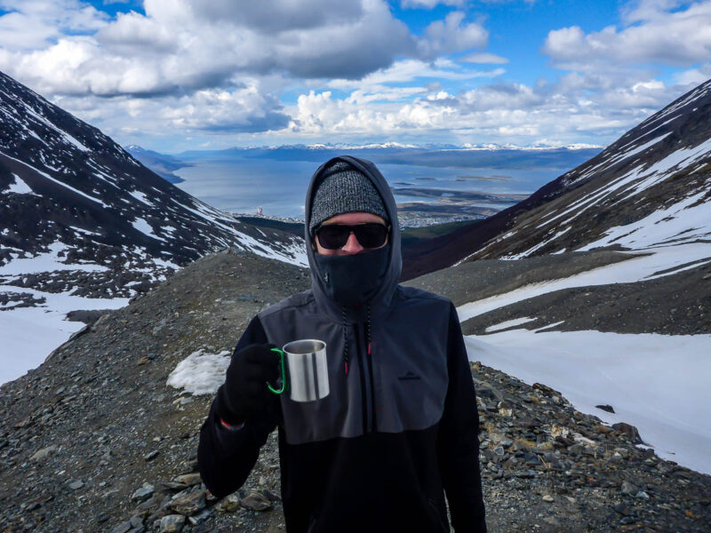 A man holding up a camping mug overlooking snow capped mountains and a body of water in the distance