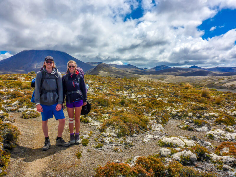 A man and woman smiling at the camera while hiking with bush, dirt, and rocks around them, mountains in the background and clouds in the sky