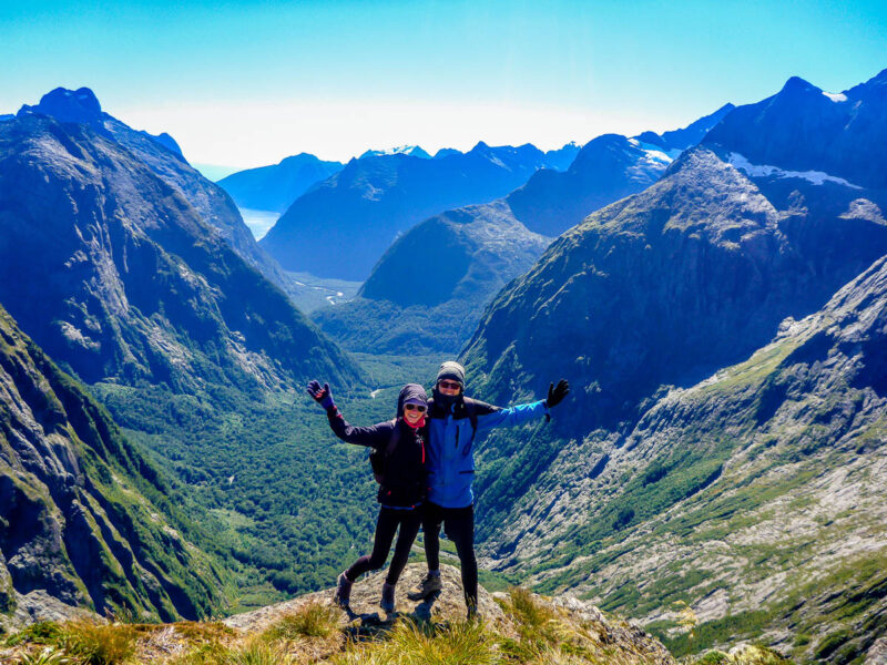 A woman and man with their arms outstretched on the side on a mountain with rolling hills and forest covered mountains behind them and blue sky