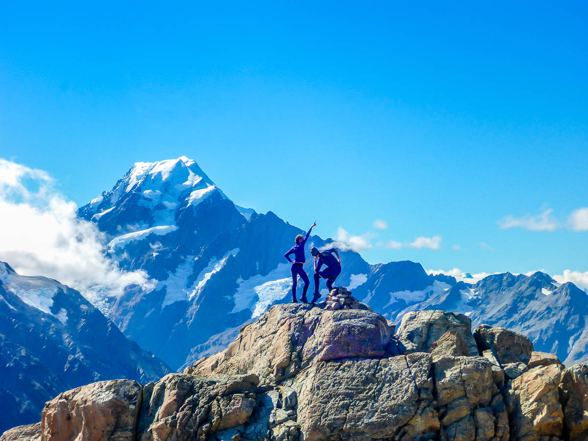 A woman and man posing on top of a mountain of large boulders with a snow capped mountain in the distance