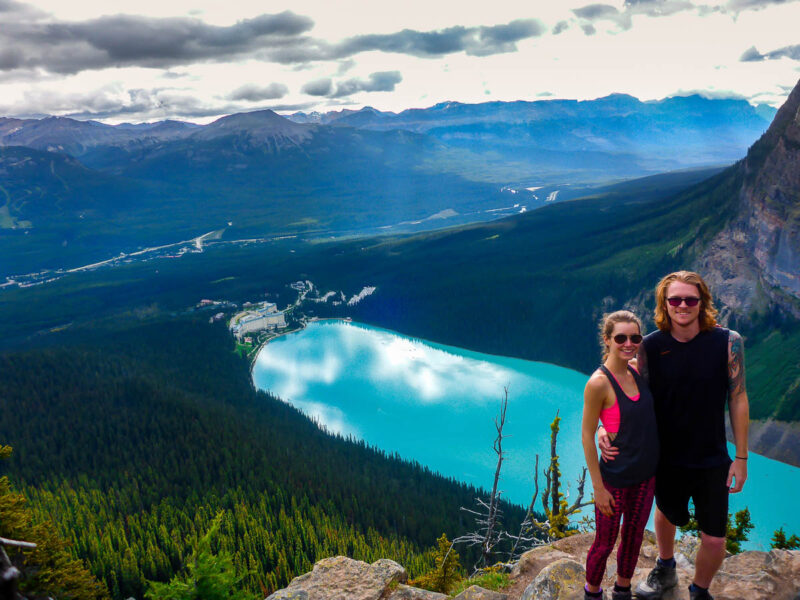 A woman and man posing on the side of a mountain with a turquoise colored lake down below, green forests, and mountains all around. Big Beehive Hike, Lake Louise, Alberta, Canada