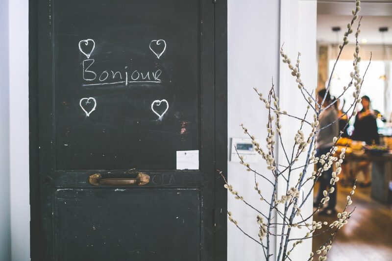 A black chalkboard door with the word "Bonjour" and four hearts drawn on it with a small plant and blurry cafe scene in the background