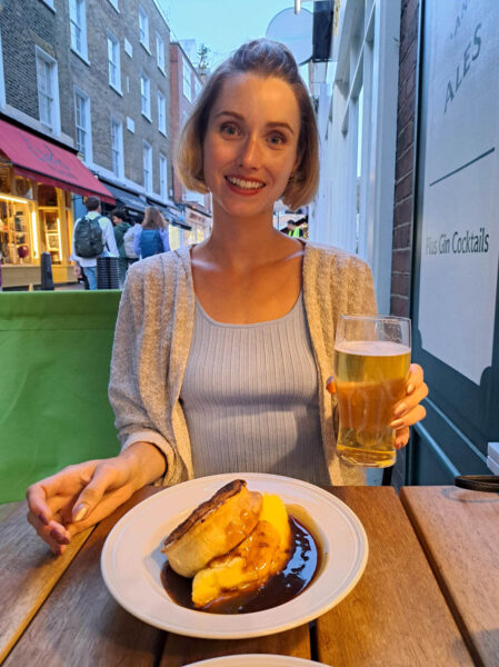 A woman sitting and smiling for the camera holding a beer. In front of her is a meal of a vegan pie and mashed potatoes