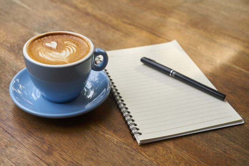 A cup of coffee on a saucer beside a blank notebook and pen on a wooden table