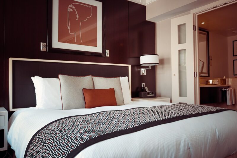 Hotel bed with 5 decorative pillows, a throw at the end of the bed, a burgundy colored headboard, side table and lamps, burgundy wall with artwork