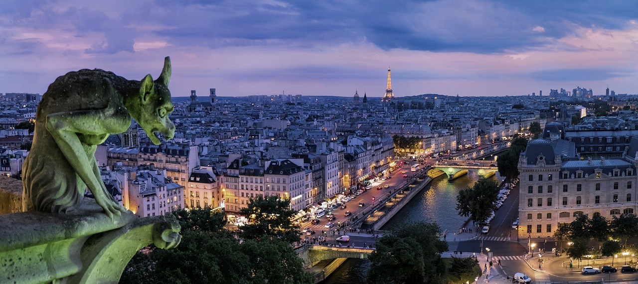 Paris city at night with a gargoyle looming on the side of a building and the lit up Eiffel Tower in the distance