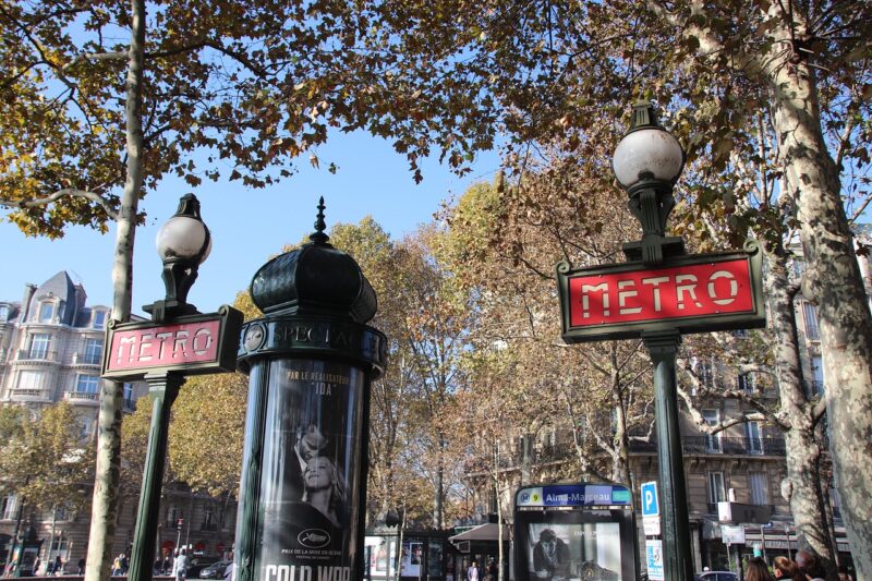 Two red Metro signs in Paris with tree leaves hanging above and city scene in the background