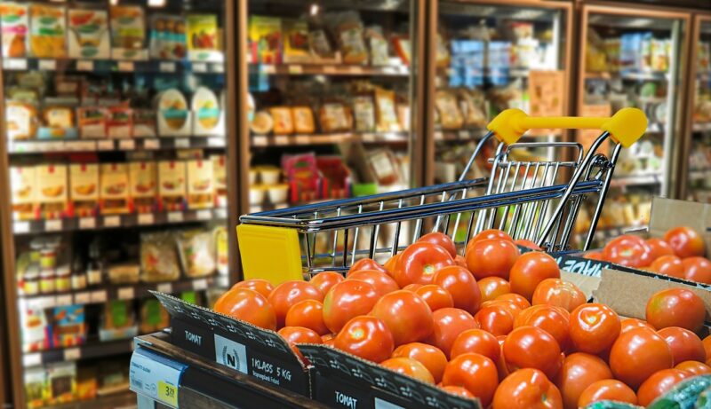 Tomatos in a grocery store with a shopping cart beside them and refrigerators full of products behind them