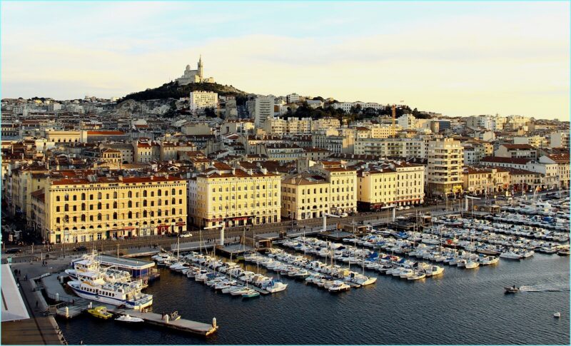 Marseille's Vieux Port at sunset with boats docked, waterfront buildings, and the Notre Dame Basilica on a hill in the background