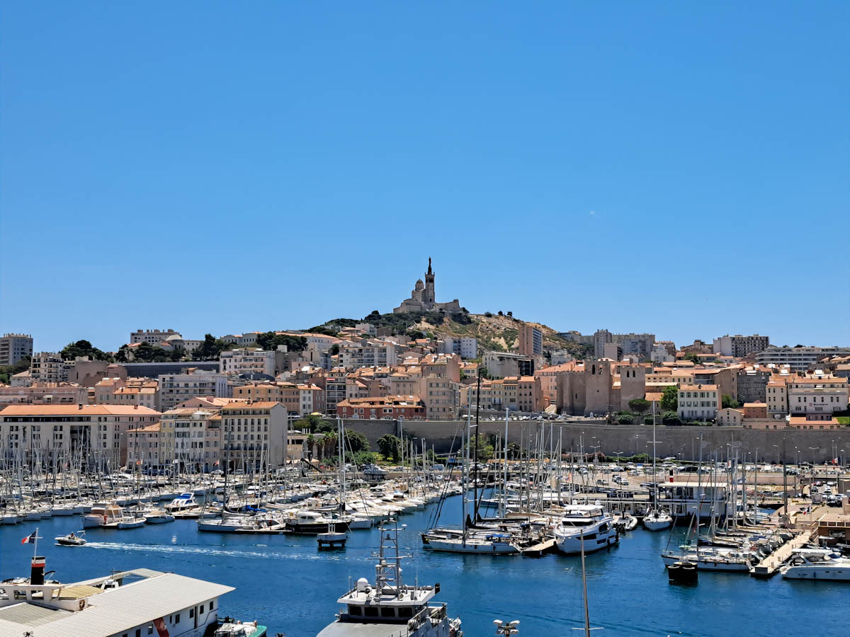 View of Marseille Vieux Port with sailing boats, speed boats and more in the marina. Cityscape behind with the Notre Dame de la Garde atop a hill in the background.