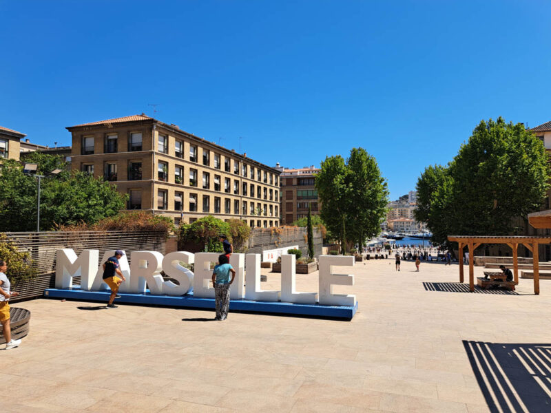 Marseille sign with buildings behind it, blue skies, and the Vieux Port in the background with spotless streets, despite the travel myth that Marseille is dirty.