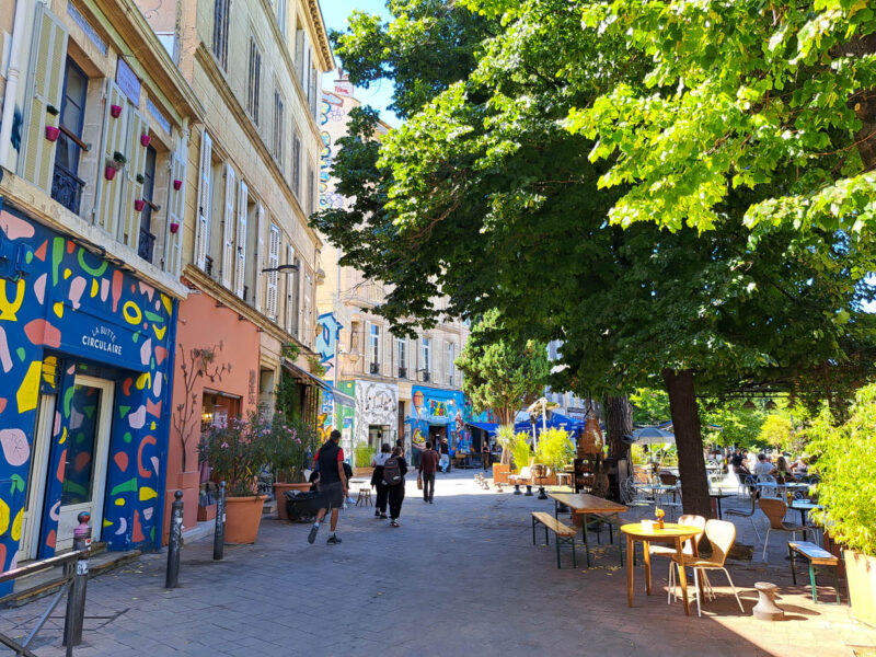 A view of a street in the Cours Julien neighborhood in Marseille with colorful street art lined business on one side and bright green trees on the other.