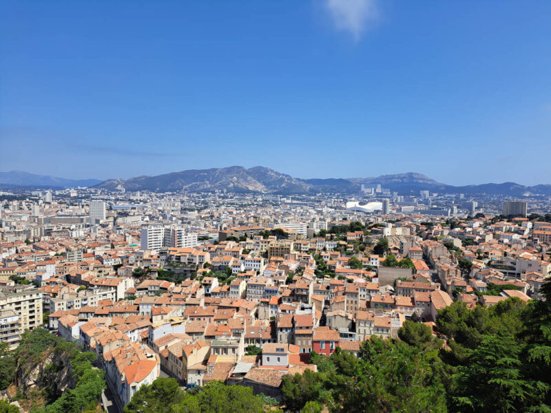 View of Marseille from the top of a hill with buildings, houses, trees and mountains in the background.